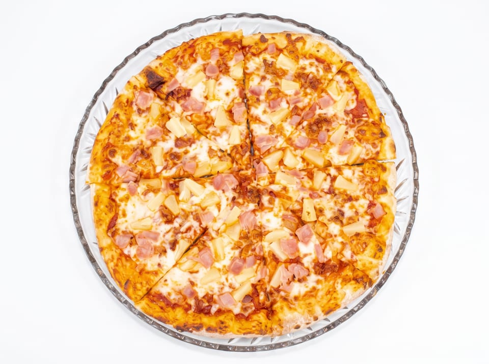 Overhead view of a whole Hawaiian pizza with ham and pineapple on a clear glass plate.