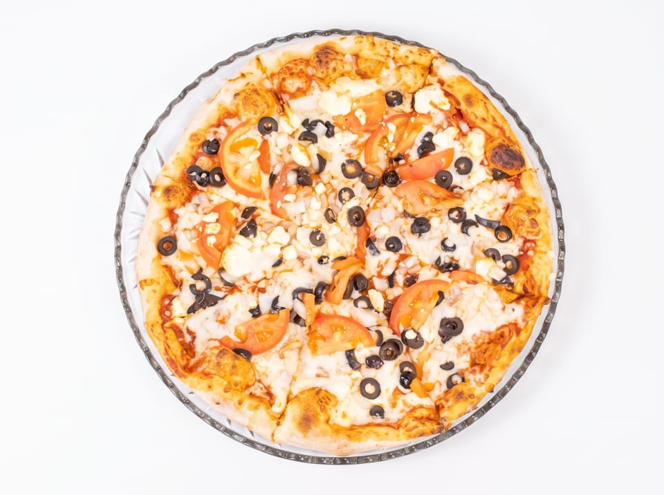 A whole pizza with tomato slices, black olives, and cheese on a clear glass plate.