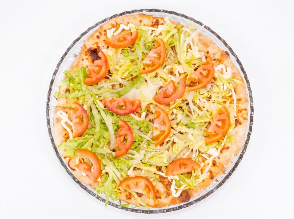 Overhead view of a pizza topped with shredded lettuce, tomato slices, and white sauce on a clear plate.